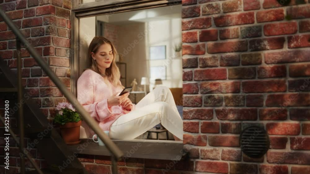 Poster a beautiful woman in comfortable casual clothes using smartphone while sitting on a windowsill on a 
