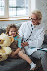 Kind female pediatrician doctor visiting his patient at home, examining little girl, writing prescription. Concept of kid's health check. Measure temperature, heart beat