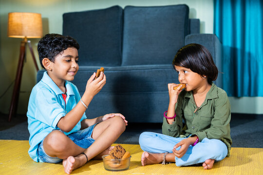 Young Sibling Kids Having Snacks Or Biscuit While Sitting On Floor At Home By Looking Around After School - Concepts Of Healthy Eating Togetherness And Childhood Lifestyle.