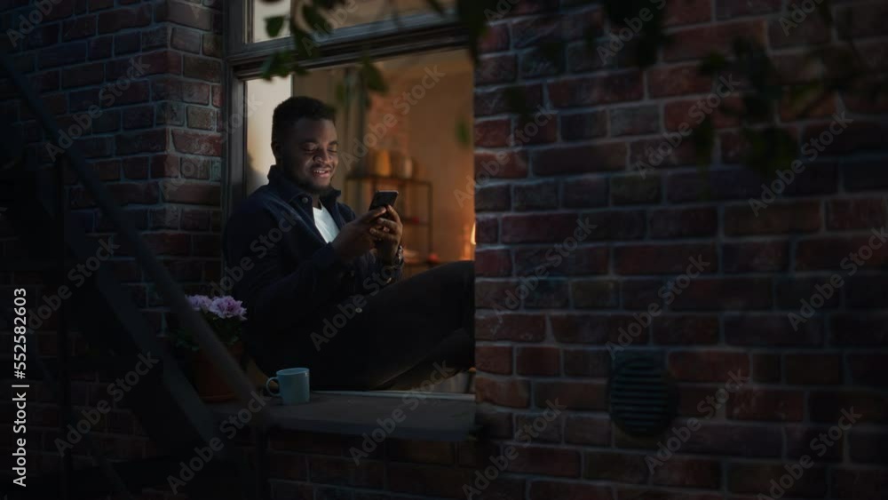 Aufkleber black man using smartphone for online shopping while sitting on his windowsill at night. e-commerce 