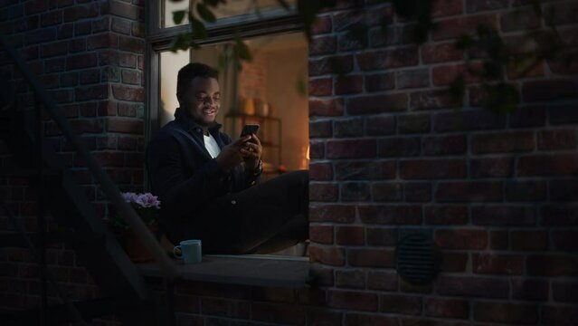 Black Man Using Smartphone for Online Shopping While Sitting on his Windowsill at Night. e-Commerce Concept of Easy Purchasing, Buying, and Ordering on Website and the Internet.