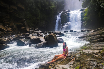 beautiful girl in bikini sits on rocks admiring powerful nauyaca waterfall in Costa Rica, large...