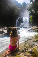 beautiful girl in bikini sits on rocks admiring powerful nauyaca waterfall in Costa Rica, large tropical waterfall in Costa Rican rainforest