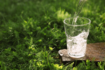 Pouring fresh water into glass on stone in green grass outdoors. Space for text