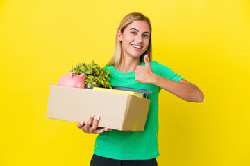 Young Uruguayan girl making a move while picking up a box full of things isolated on yellow background giving a thumbs up gesture