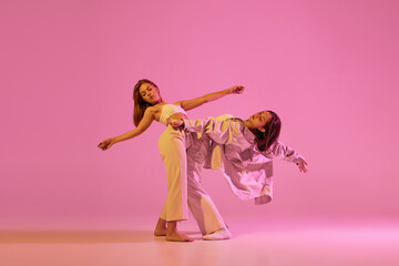 Young girls, couple of dancers in sports style clothes dancing experimental dances isolated over light pink background in neon. Concept of music, emotions, dance