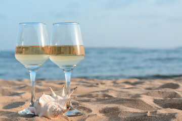 Glasses of tasty wine and seashell on sand near sea, space for text