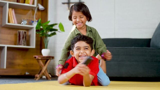 Happy smiling siblings kids fighting for remote control while watching tv or television at home - concept of childhood lifestyle, siblings quarrel and technology.