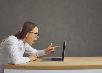 Side view of emotional woman with puzzled expression sitting in front of laptop and pointing at screen. Young woman looks amazed at the shocking content sitting on the background of a gray wall.