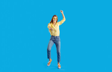 Obraz na płótnie Canvas Happy young woman dancing feeling euphoric and celebrating success on blue background. Joyful excited casual girl having fun celebrating and rejoicing in good news. Concept of good luck. Full length.