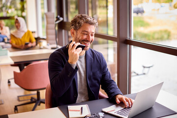 Mature Businessman Working On Laptop At Desk In Office Talking Into Mic Of Mobile Phone