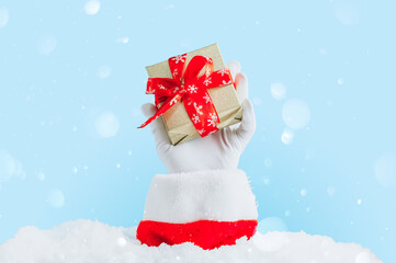 Santa's hand sticking out of a snowdrift. Santa Claus gloved hands holding gift box against light blue bokeh background.