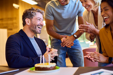 Staff Celebrating Birthday Of Male Colleague At Desk In Office With Cake