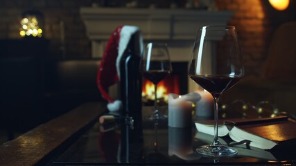 Glass of red wine on table at home at Christmas and wine bottle with Santa hat. Winter mood with fireplace in cosy dark living room.