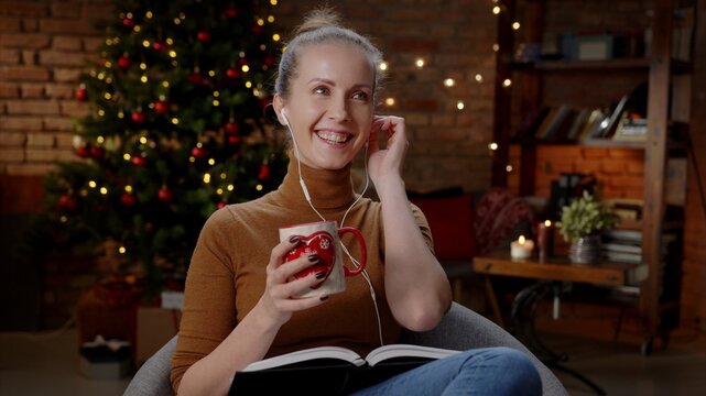 Adult woman reading book listening to music at home at Christmas in winter, dark room with Christmas tree.
