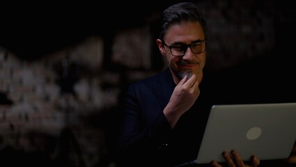 Business portrait - happy smiling businessman in loft office holding laptop computer. Mature age, middle age, mid adult man in 50s, white caucasian, Low light, dark copy space.