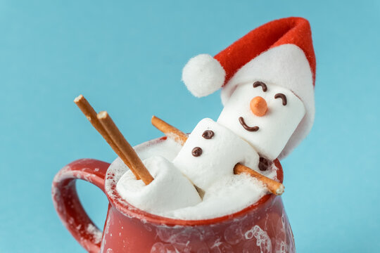 Marshmallow snowman taking hot tub in a red ceramic cup full of cocoa with milk foam. Christmas holidays blue background. Wintertime concept. Hot chocolate with marshmallow and festive decoration.