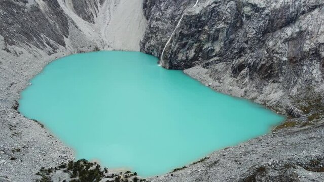 Drone flyover of Laguna 69 (lake 69) or Laguna Sesentinueve, a luminescent green turquoise chemical lake in Peru. Camera flies forwards low over the lake with the glacial waterfall in the background.