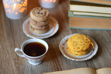 Plates with various cookies, cups of tea or coffee, vintage books, reading glasses and lit candles on the table. Selective focus.