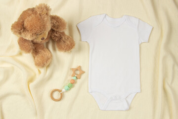 White cotton baby short sleeve bodysuit, teddy bear and natural wooden toy on beige blanket throw...