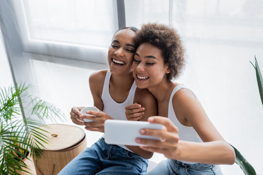 cheerful african american lesbian woman taking selfie with girlfriend holding cup of coffee.