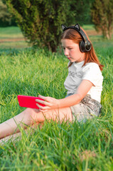 teenage girl sits in a park on the grass with a smartphone in her hands. 12 year old girl listening to music in headphones sitting in the park