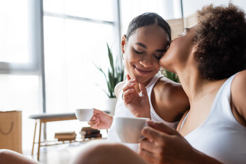 lesbian african american woman kissing cheek of smiling girlfriend with cup of coffee.