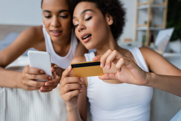 amazed african american lesbian woman holding credit card near girlfriend with smartphone.
