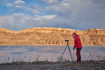 senior male photographer with a camera on tripod on a shore of frozen lake in Colorado foothills
