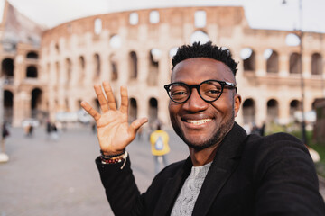 a black boy with eyeglasses on holiday at the colosseum taking a selfi smiling.