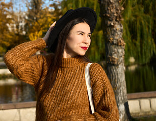 Selective focus of smiling fashionable young tanned woman in hat among autumn foliage outdoors in city park autumn. Positive emotion concept.