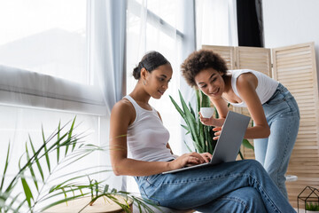 young african american lesbian woman using laptop near curly girlfriend holding cup of coffee.