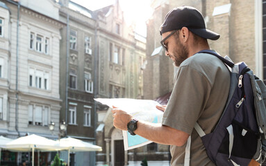 Man tourist in cap and sunglasses traveling around old town in Europe using paper map.