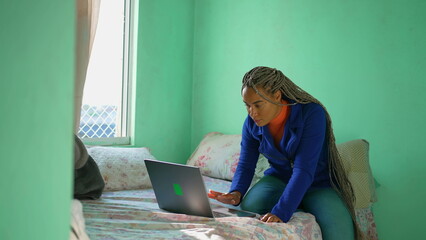 A young Brazilian adult girl sitting in bed opening laptop screen. African American millennial...