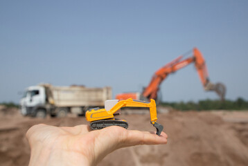 On the arm is a small orange toy excavator. Behind him, out of focus, is a real working excavator...