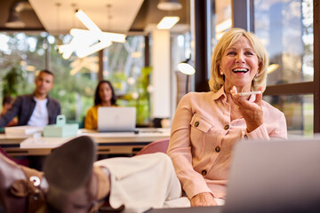 Mature Businesswoman With Feet On Desk Working On Laptop In Office Talking Into Mic Of Mobile Phone