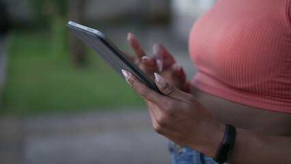 One young black woman using tablet outdoors. African American adult girl standing outside leaning on wall holding device touching screen