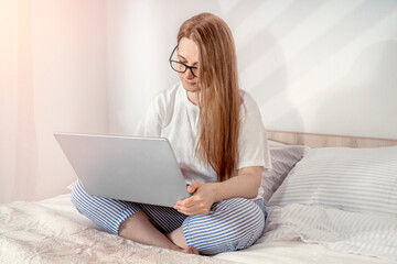 Woman working with laptop in bed at home or hotel in the morning. Lady relaxing in a nightgown. Girl wearing nightie on clean white bedding, cozy blanket