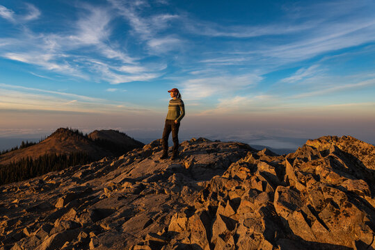 Woman standing on mountain summit looking out with sunset light and hazy in the background under blue skies