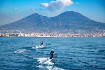 Two people ride an electric surfboard in the Gulf of Naples, Italy, against the backdrop of Mount...