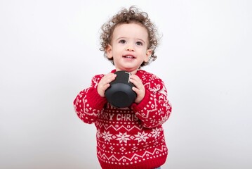 Beautiful little baby boy with curly hair wearing red Christmas knitted sweater against white background holding black wireless speaker listening to music and jingle songs. 