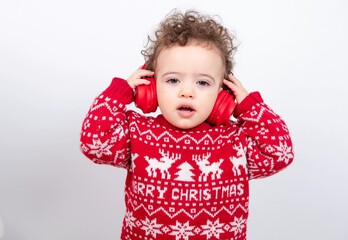 Beautiful little baby boy with curly hair wearing Christmas knitted sweater against white background wearing red headphones listening to music and looking aside. 