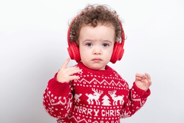 Beautiful little baby boy with curly hair wearing red Christmas knitted sweater against white background enjoys listening to videos or music wearing red headphones, clapping and dancing. 