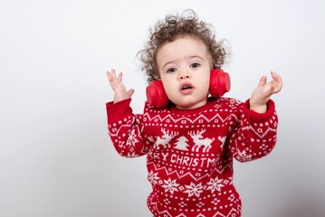 Beautiful little baby boy with curly hair wearing red Christmas knitted sweater against white background wearing red wireless headphones listening to music and dancing. 