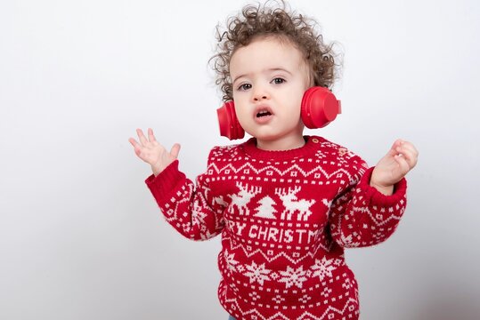 Beautiful little baby boy with curly hair wearing red Christmas knitted sweater against white background wearing red wireless headphones listening to music and dancing. 