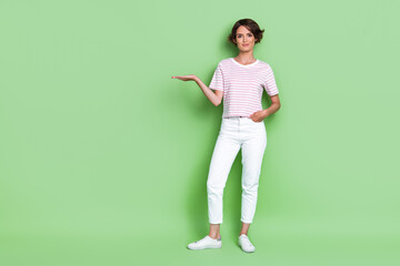 Full body photo of nice young woman hold palm empty space new product promo dressed trendy striped look isolated on green color background