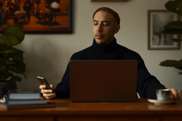 A caucasian man wearing a orange beanie and a blue knitted sweatshirt sitting at a wooden table...