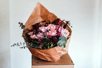 Un ramo de flores en tonos rosas y granates envuelto en papel marrón dentro de una caja de...