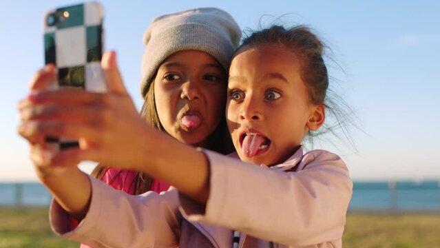 Children, friends and phone selfie at beach park while making silly faces with tongue out. Kids, mobile and girls taking pictures on 5g smartphone for profile picture, happy memory or social media.