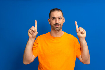 Portrait of a happy bearded hispanic man in orange t-shirt pointing with index fingers up where copyspace is isolated over blue background.